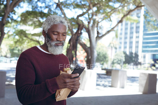 Uomo anziano afroamericano in strada mangiare panino e utilizzando smartphone. nomade digitale in giro per la città. — Foto stock