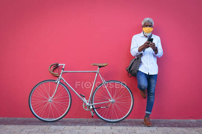 Африканский пожилой человек в маске стоит рядом с велосипедом на улице, используя смартфон. Цифровой кочевник в городе во время пандемии коронавируса. — стоковое фото