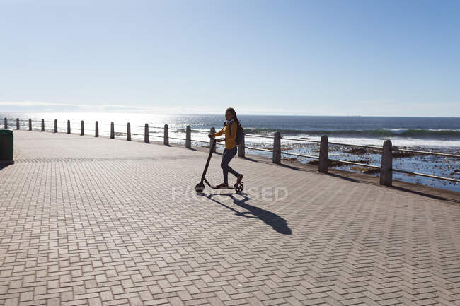 Happy african american woman wearing headphones and backpack riding scooter on promenade by the sea. Digital nomad on the go lifestyle. — Stock Photo
