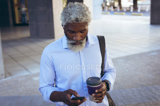 Hombre mayor afroamericano de pie en la calle sosteniendo café y usando un teléfono inteligente. nómada digital en la ciudad. - foto de stock