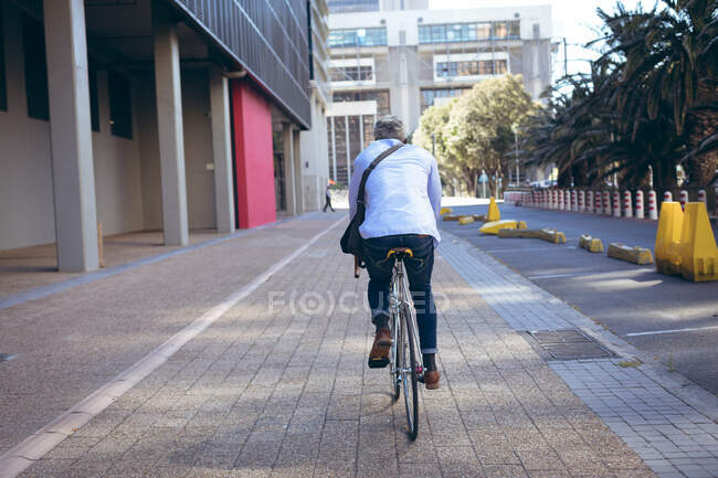 Rear view of african american senior man riding bicycle in the street. digital nomad out and about in the city. — Stock Photo