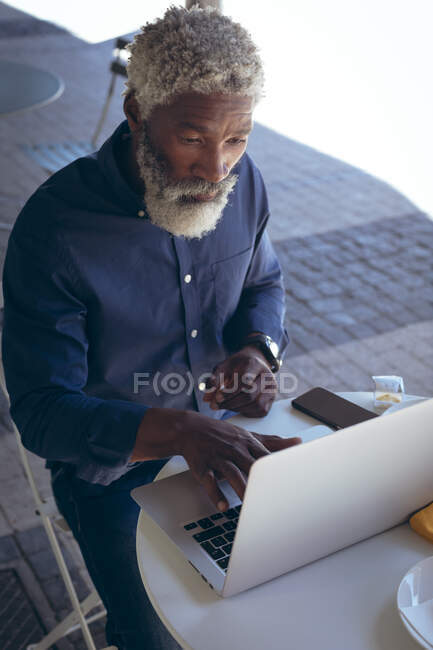 Hombre mayor afroamericano sentado en la mesa fuera de la cafetería usando un portátil. nómada digital en la ciudad. - foto de stock