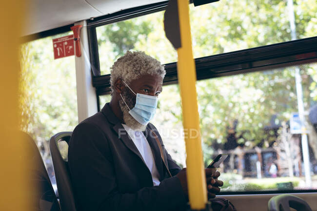 Hombre mayor afroamericano con máscara facial sentado en el autobús usando un teléfono inteligente. nómada digital en la ciudad durante la pandemia de coronavirus covid 19. - foto de stock