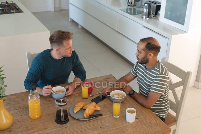 Casal masculino gay multi étnico tomando café da manhã e conversando em casa. Ficar em casa em auto-isolamento durante o bloqueio de quarentena. — Fotografia de Stock