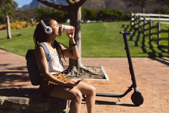 Afroamerikanerin entspannt, mit Kopfhörern sitzend und Kaffee trinkend im Park. Digitaler Nomade unterwegs. — Stockfoto