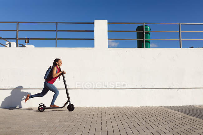 Smiling african american woman wearing backpack riding scooter on promenade by the sea. Digital nomad on the go lifestyle. — Stock Photo