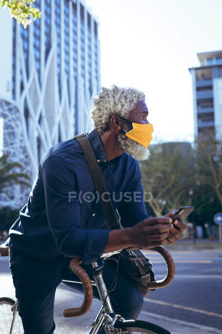 Африканский пожилой человек в маске для лица сидит на велосипеде на улице, используя смартфон. Цифровой кочевник в городе во время пандемии коронавируса. — стоковое фото