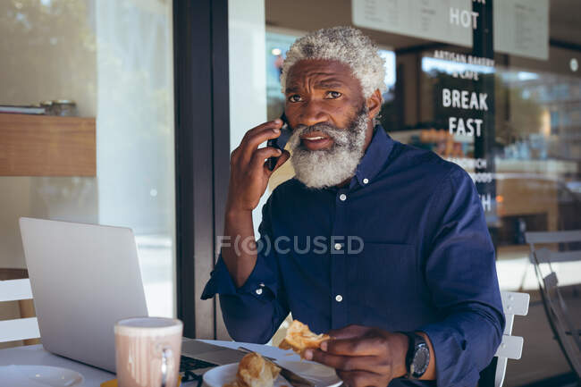 African american senior man sitting at table outside cafe talking on smartphone. digital nomad out and about in the city. — Stock Photo