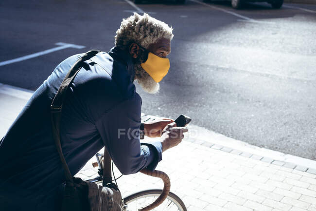 Ein afroamerikanischer älterer Mann mit Gesichtsmaske lehnt auf der Straße mit dem Smartphone auf einem Fahrrad. digitaler Nomade unterwegs in der Stadt während Coronavirus covid 19 Pandemie. — Stockfoto