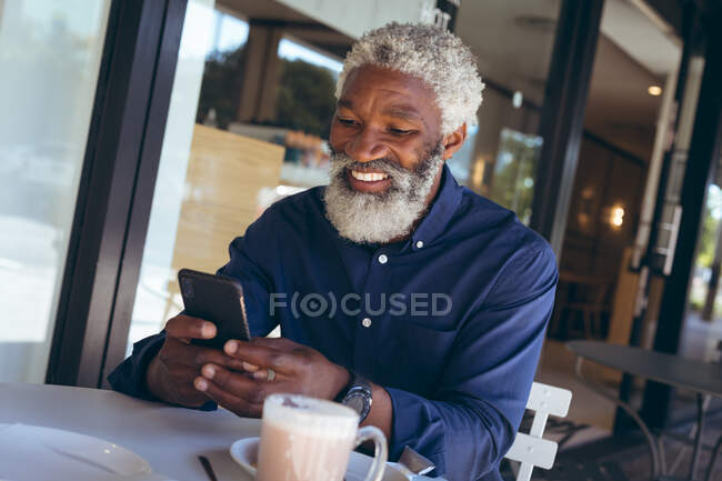 Африканский старший американец сидит за столом возле кафе с кофе, используя смартфон и улыбаясь. цифровая реклама в городе. — стоковое фото