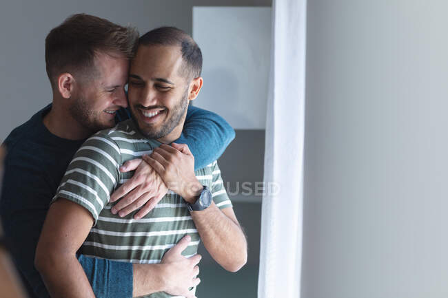 Multi-ethnische schwule männliche Pärchen lächeln, stehen am Fenster und umarmen sich zu Hause. Genießen Sie die Zeit zu Hause in Selbstisolierung während der Quarantäne Lockdown. — Stockfoto