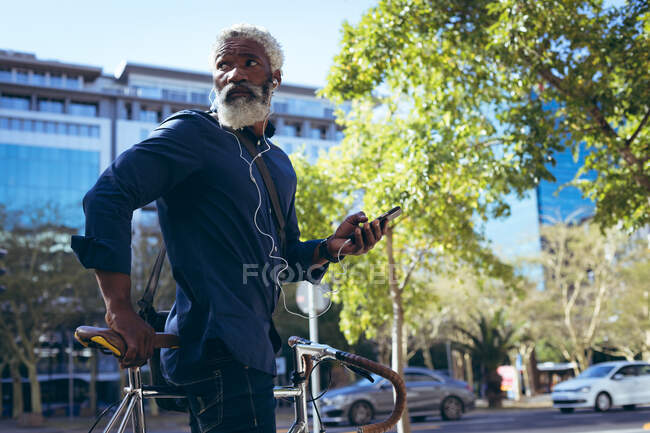 Ein afroamerikanischer älterer Mann mit Kopfhörern sitzt auf einem Fahrrad auf der Straße und benutzt ein Smartphone. digitaler Nomade in der Stadt unterwegs. — Stockfoto