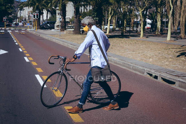 Afro-americano idoso vestindo máscara facial andando de bicicleta através da estrada em uma travessia de pedestre. nômade digital para fora e sobre na cidade durante coronavírus covid 19 pandemia. . — Fotografia de Stock