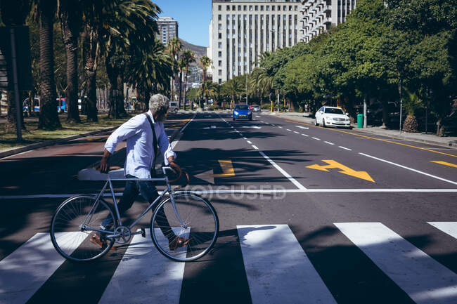 Ein afroamerikanischer Senior rast mit dem Fahrrad auf einem Fußgängerüberweg über die Straße. digitaler Nomade in der Stadt unterwegs. — Stockfoto