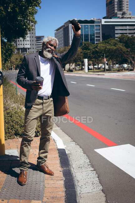 Ein älterer afroamerikanischer Mann steht mit Kaffee und Smartphone am Straßenrand und begrüßt ein Taxi. digitaler Nomade in der Stadt unterwegs. — Stockfoto