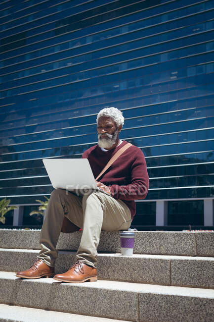 Hombre mayor afroamericano sentado en escalones fuera de un edificio con café para llevar usando un portátil. nómada digital en la ciudad. - foto de stock
