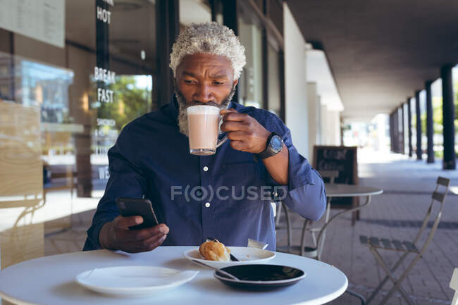 African american senior man sitting at table outside cafe drinking coffee and using smartphone. digital nomad out and about in the city. — Stock Photo