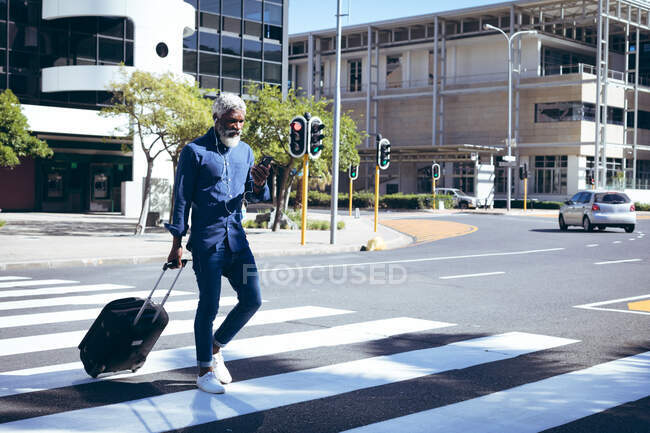 Homme âgé afro-américain portant des écouteurs et utilisant une valise à roulettes smartphone à travers la route sur un passage pour piétons. nomade numérique dans la ville. — Photo de stock