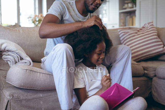 African american man sitting on bed doing his daughter hair. staying at home in self isolation during quarantine lockdown. — Stock Photo