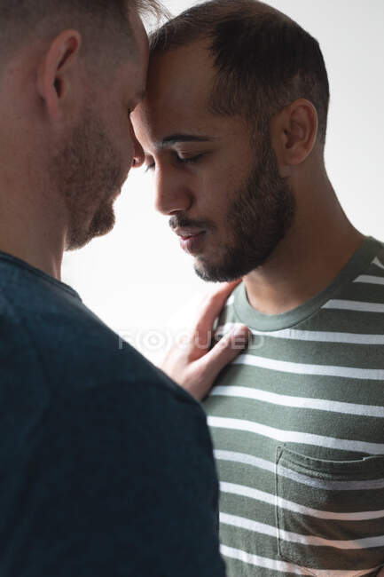 Multi ethnic gay male couple standing by window hugging at home. Staying at home in self isolation during quarantine lockdown. — Stock Photo