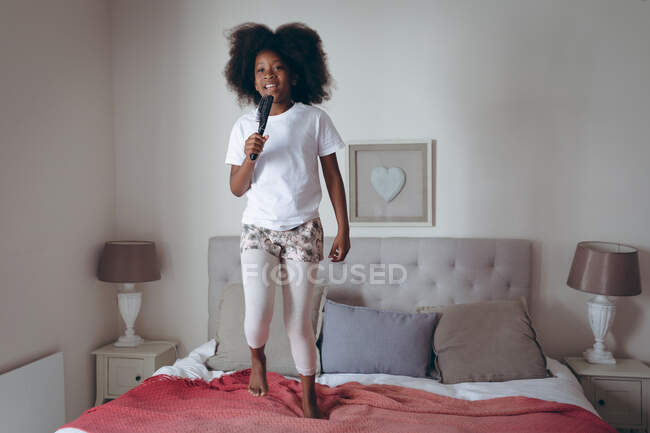 Chica afroamericana de pie en la cama sosteniendo cepillo de pelo fingiendo cantar. permanecer en casa en aislamiento durante el bloqueo de cuarentena. - foto de stock