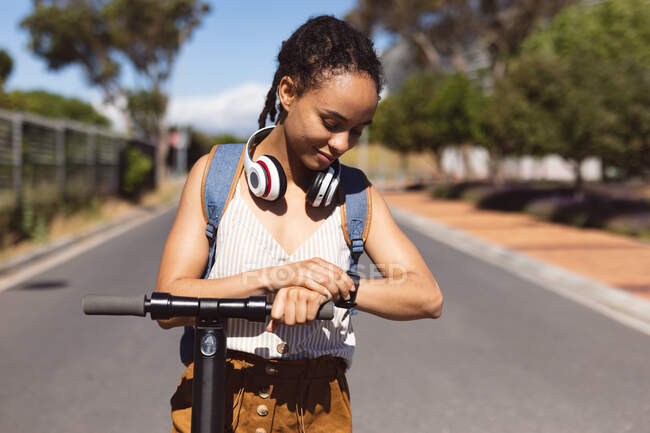 Smiling african american woman wearing headphones standing on scooter using smartwatch in street. Digital nomad on the go lifestyle. — Stock Photo