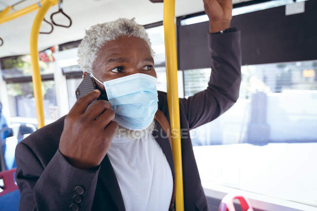 Африканский пожилой человек в маске, стоящий в автобусе и разговаривающий по смартфону. Цифровой кочевник в городе во время пандемии коронавируса. — стоковое фото