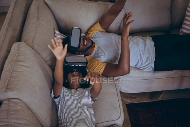 Hombre afroamericano y su hija tumbados en la sala de estar con auriculares vr. permanecer en casa en aislamiento durante el bloqueo de cuarentena. - foto de stock
