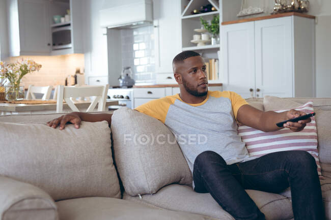 Homem afro-americano sentado na sala de estar a ver televisão. ficar em casa em auto-isolamento durante o confinamento de quarentena. — Fotografia de Stock