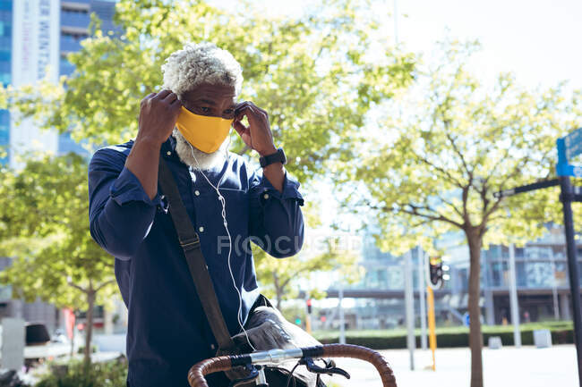 African american senior man wearing face mask sitting on bicycle in street putting on earphones. digital nomad out and about in the city during coronavirus covid 19 pandemic. — Stock Photo