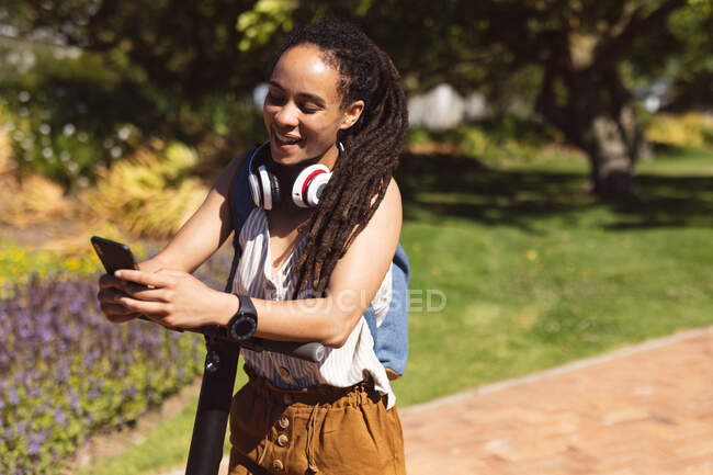Donna afroamericana sorridente che indossa cuffie in piedi su scooter utilizzando smartphone in strada. Nomade digitale in movimento lifestyle. — Foto stock