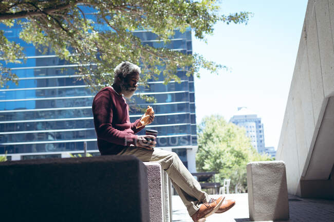 Ein älterer afroamerikanischer Mann sitzt auf der Straße, isst Sandwich und hält Kaffee. digitaler Nomade in der Stadt unterwegs. — Stockfoto