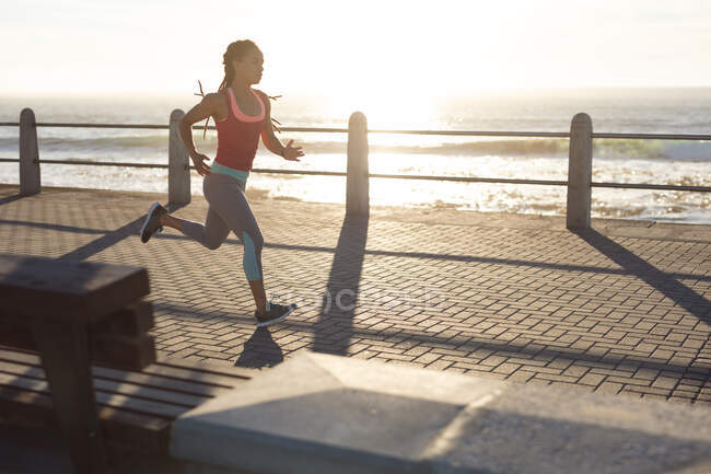 African american woman exercising on a promenade by the sea jogging. fitness healthy outdoor lifestyle. — Stock Photo