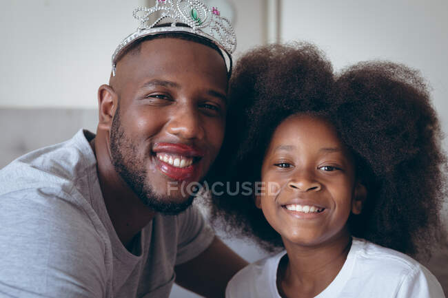 African american man wearing tiara having makeup put on by his daughter. staying at home in self isolation during quarantine lockdown. — Stock Photo