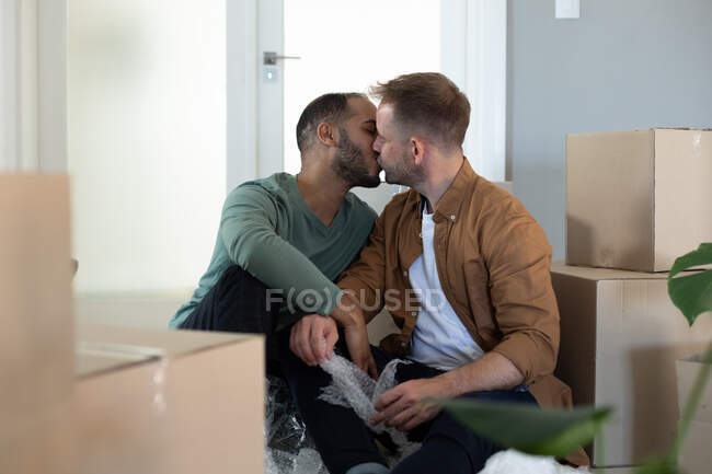 Happy multi ethnic gay male couple sitting surrounded by boxes and kissing at home. Staying at home in self isolation during quarantine lockdown. — Stock Photo