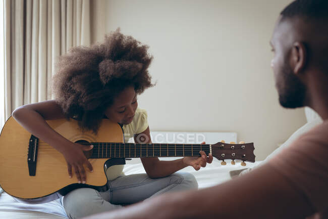 Homem afro-americano e sua filha sentados na cama menina está tocando guitarra. ficar em casa em auto-isolamento durante o confinamento de quarentena. — Fotografia de Stock