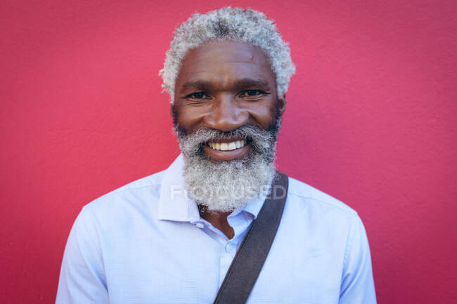 Ritratto di anziano afroamericano con la barba in piedi contro il muro rosso per strada sorridente. nomade digitale in giro per la città. — Foto stock