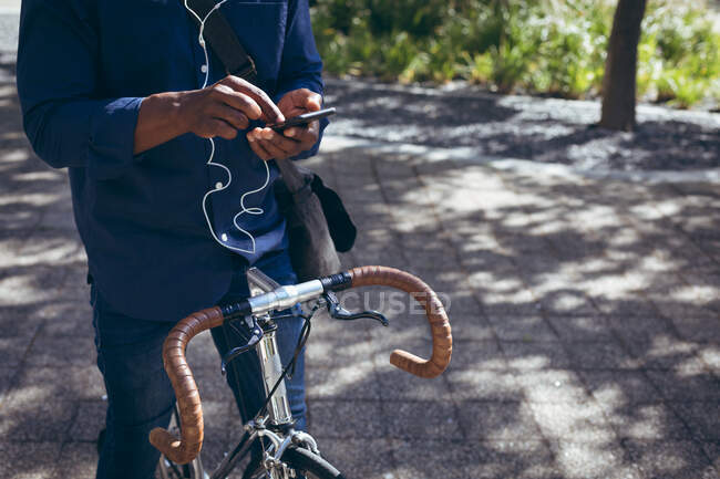 Midsection of african american senior man wearing earphones sitting on bike in street using smartphone. nomade numérique dans la ville. — Photo de stock
