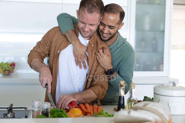Casal masculino gay multi étnico sorrindo, preparando comida e abraçando em casa. Ficar em casa em auto-isolamento durante o bloqueio de quarentena. — Fotografia de Stock