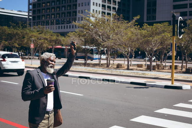 African american senior man standing at roadside holding coffee and smartphone hailing a taxi. digital nomad out and about in the city. — Stock Photo