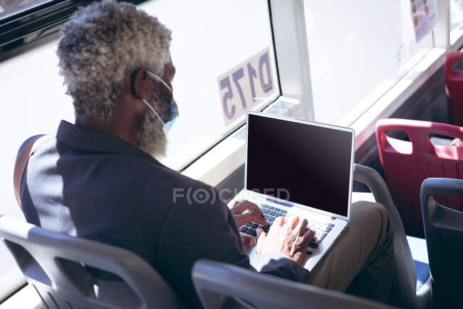 Homme âgé afro-américain portant un masque assis dans le bus à l'aide d'un ordinateur portable. numérique nomade dehors et environ dans la ville pendant coronavirus covid 19 pandémie. — Photo de stock