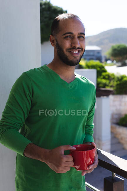 Retrato de hombre de raza mixta sonriente bebiendo taza de café en el balcón bajo el sol. Permanecer en casa en aislamiento durante el bloqueo de cuarentena. - foto de stock