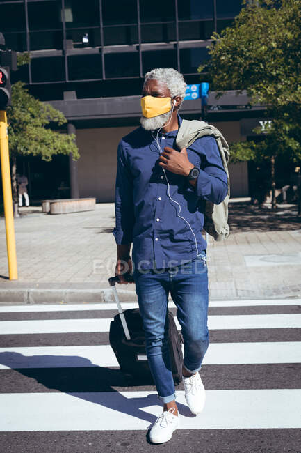 African american senior man wearing face mask and earphones wheeling suitcase across road on pedestrian crossing. digital nomad out and about in the city during coronavirus covid 19 pandemic. — Stock Photo