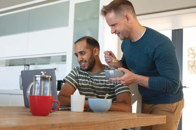 Multi pareja gay masculina étnica sonriendo y desayunando y usando el portátil en casa. Permanecer en casa en aislamiento durante el bloqueo de cuarentena. - foto de stock