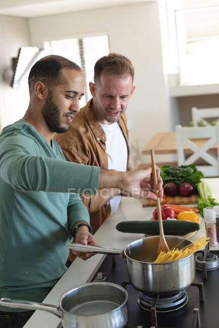 Pareja masculina gay multiétnica sonriendo y preparando comida juntos en casa. Permanecer en casa en aislamiento durante el bloqueo de cuarentena. - foto de stock