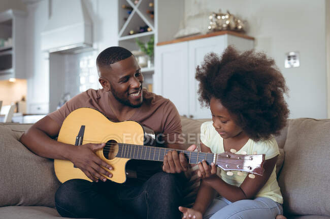 L'uomo afroamericano e sua figlia seduti sul divano suonano la chitarra. stare a casa in isolamento durante l'isolamento in quarantena. — Foto stock
