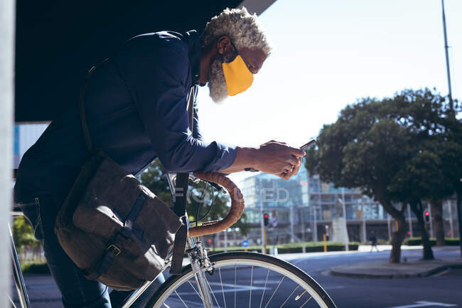 Африканский пожилой человек в маске для лица, опирающийся на велосипед на улице, используя смартфон. Цифровой кочевник в городе во время пандемии коронавируса. — стоковое фото