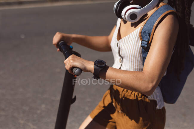 Midsection of woman wearing headphones and backpack riding scooter in park. Digital nomad on the go lifestyle. — Stock Photo