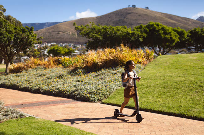 Счастливая африканская американка в рюкзаке, гуляющая со скутером в парке. Цифровой кочевник на ходу. — стоковое фото