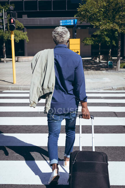 Rear view of african american senior man wearing face mask wheeling suitcase across road on pedestrian crossing. digital nomad out and about in the city during coronavirus covid 19 pandemic. — Stock Photo